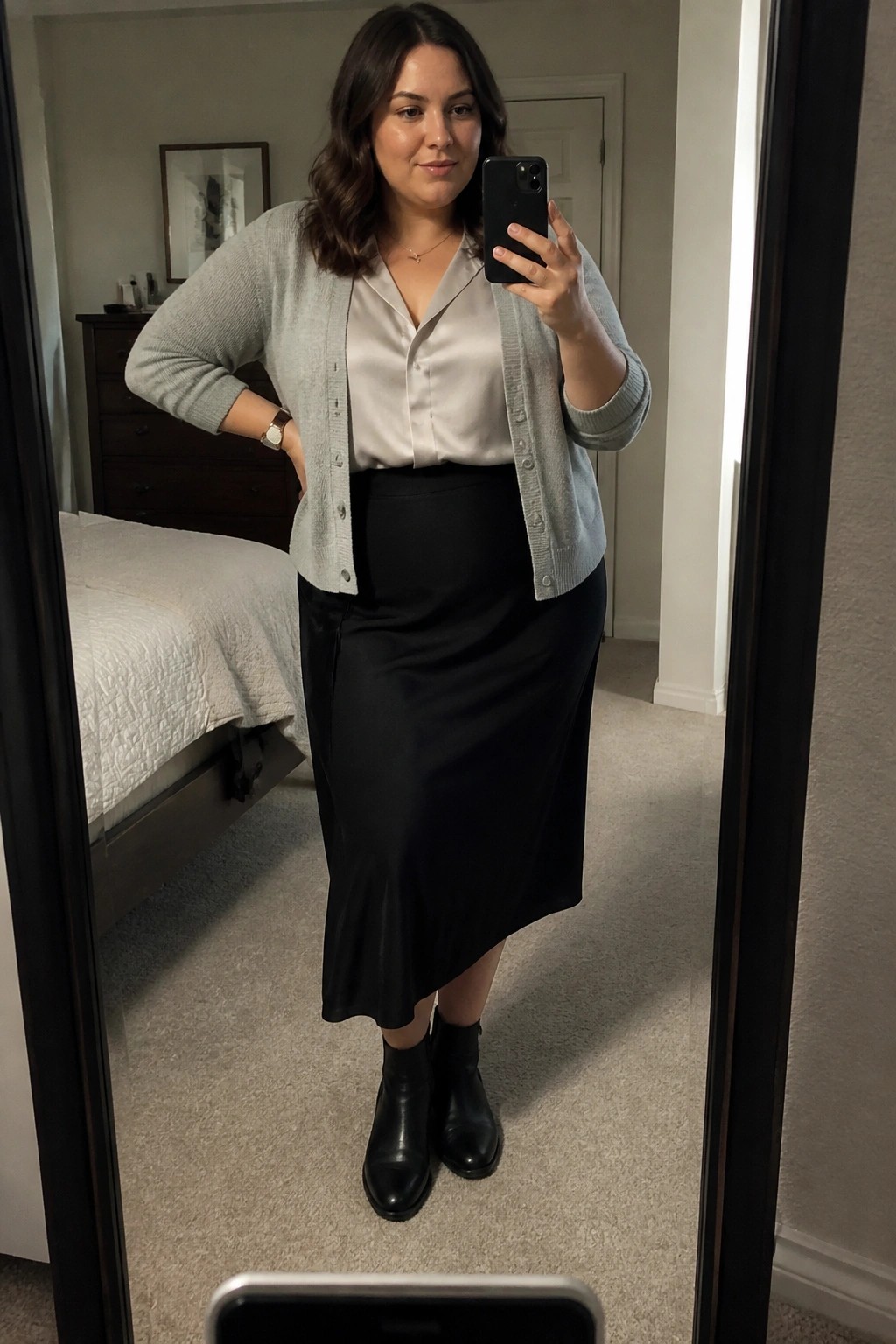 Woman in gray cardigan over white blouse and black midi skirt taking mirror selfie