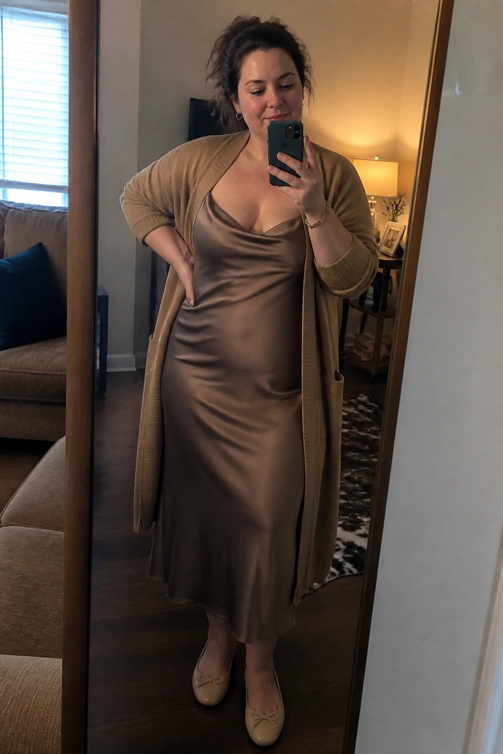 Woman in taupe satin slip dress and beige cardigan taking mirror selfie in living room