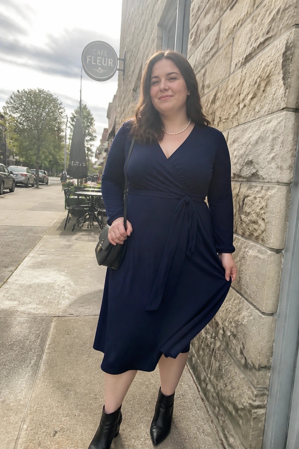 Woman in navy wrap dress holding handbag against stone wall on sidewalk