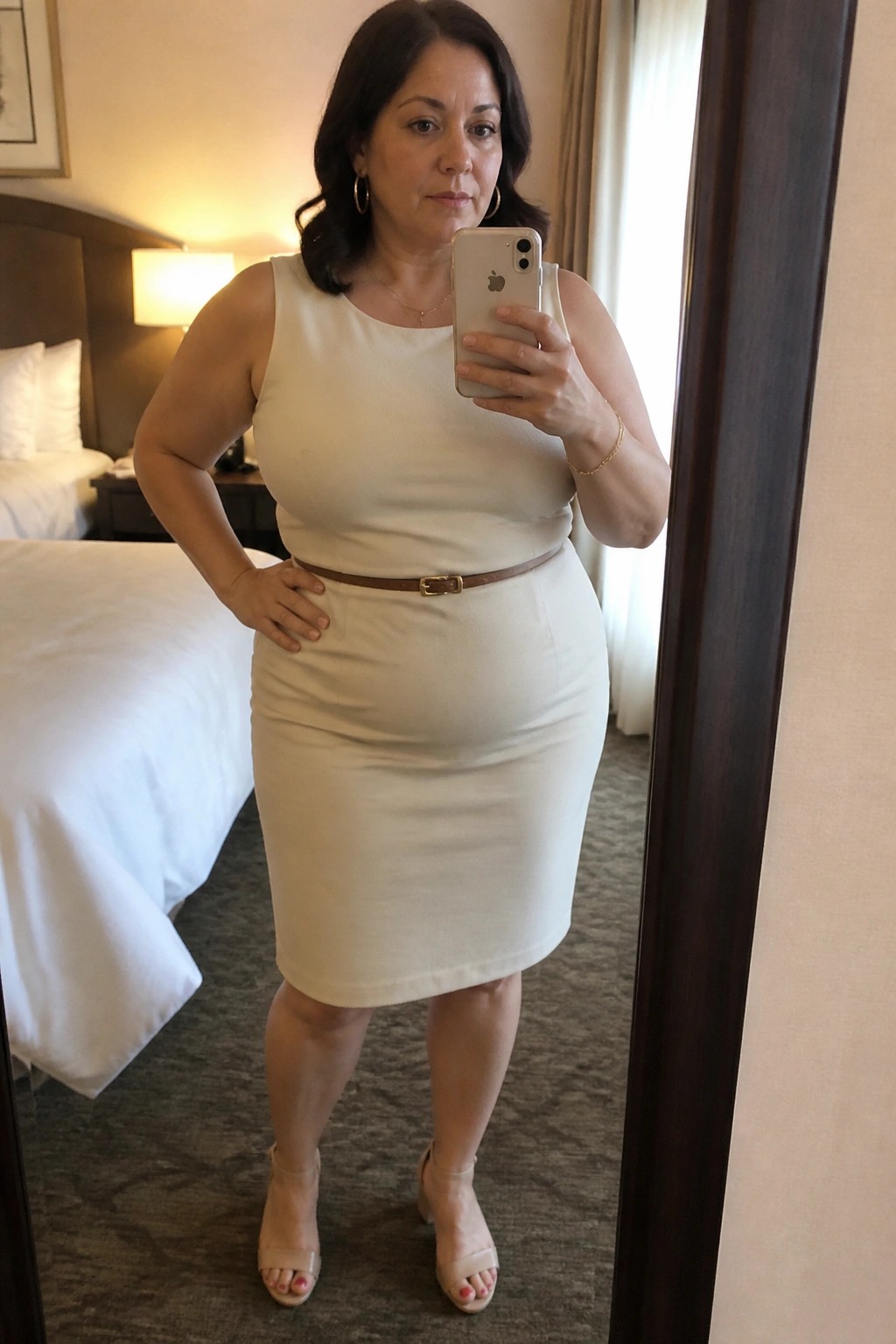 Woman in cream dress taking mirror selfie with iPhone in hotel room
