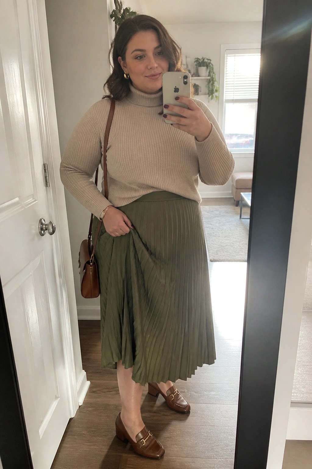 Plus size woman wearing a beige ribbed turtleneck sweater tucked into an olive green pleated midi skirt with brown leather loafers and a matching crossbody bag while holding a phone for a mirror selfie.
