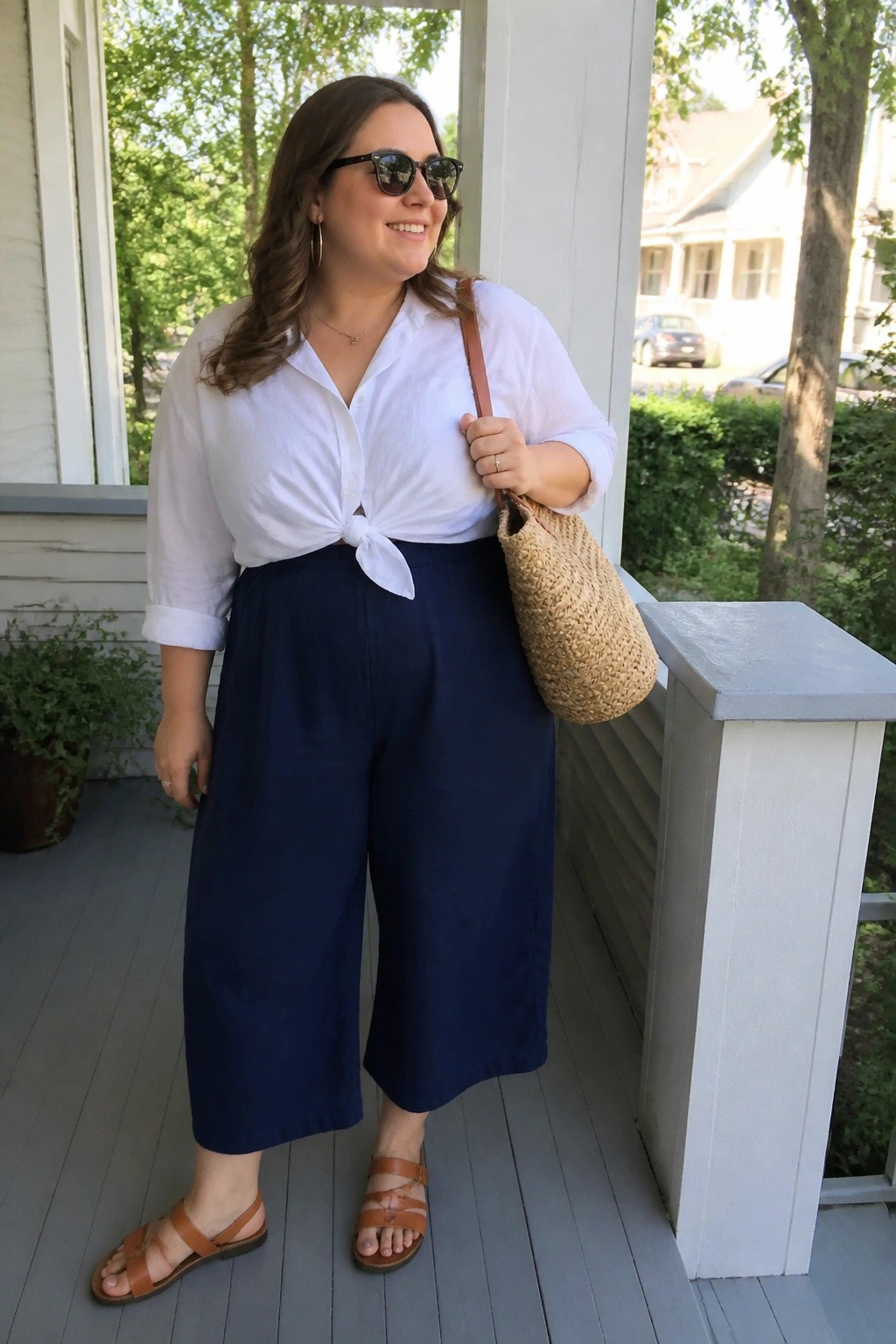 Woman in white tied shirt and navy culottes holding woven bag on porch