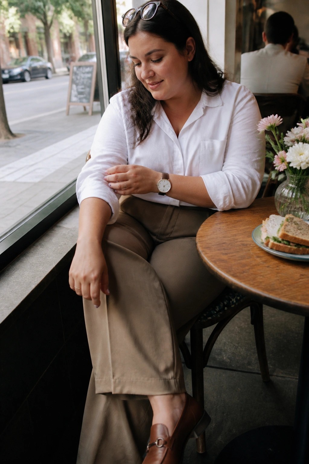 Woman in white shirt and brown pants at cafe table with sandwich and flowers