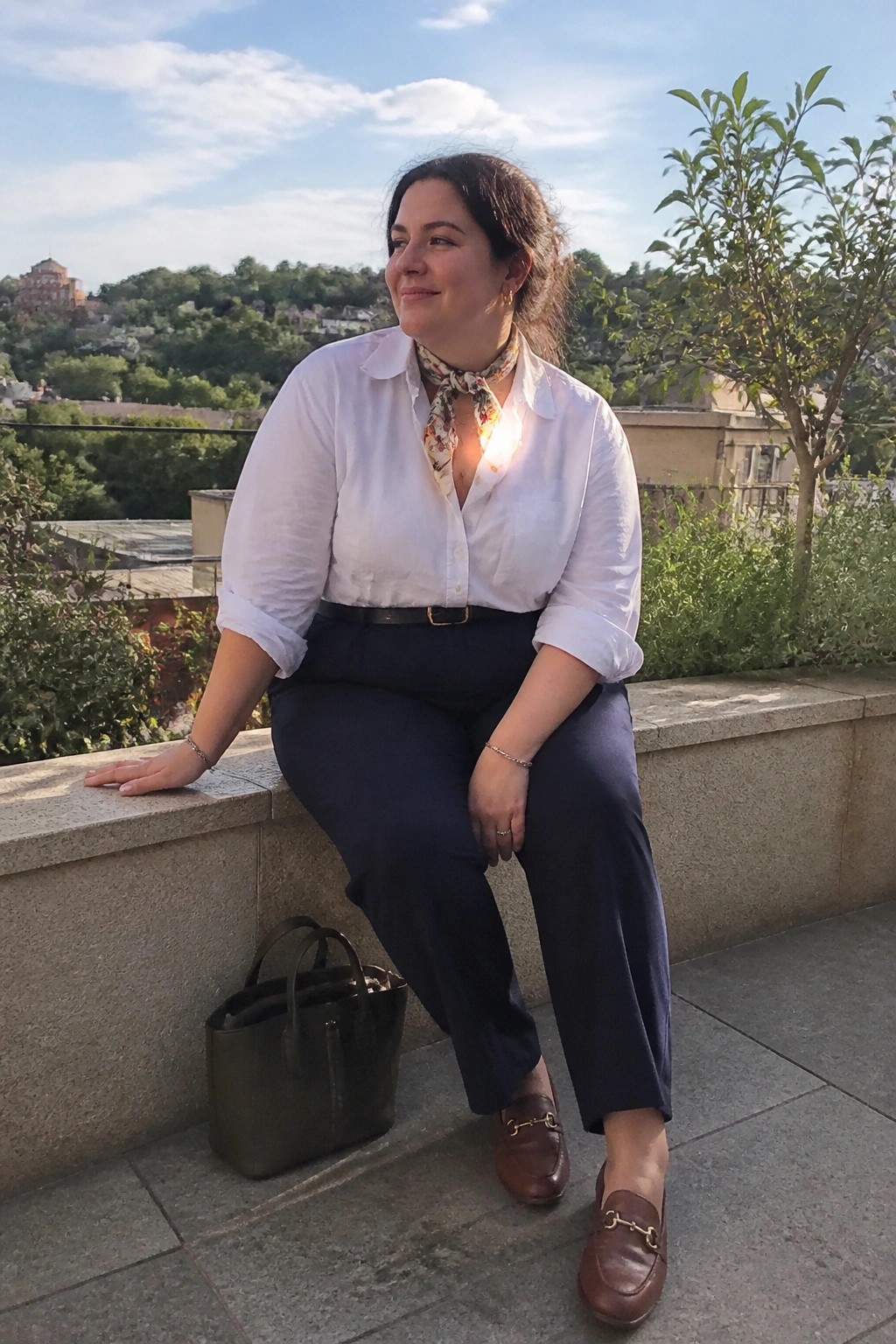 Woman in white shirt and navy pants seated on stone ledge with green bag