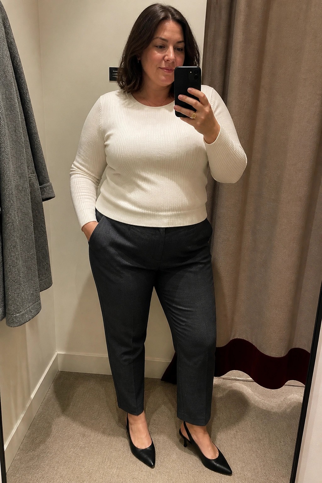 Plus size woman wearing a cream ribbed knit sweater tucked into dark gray tailored trousers with black pointed slingback heels while taking a mirror selfie in a fitting room.