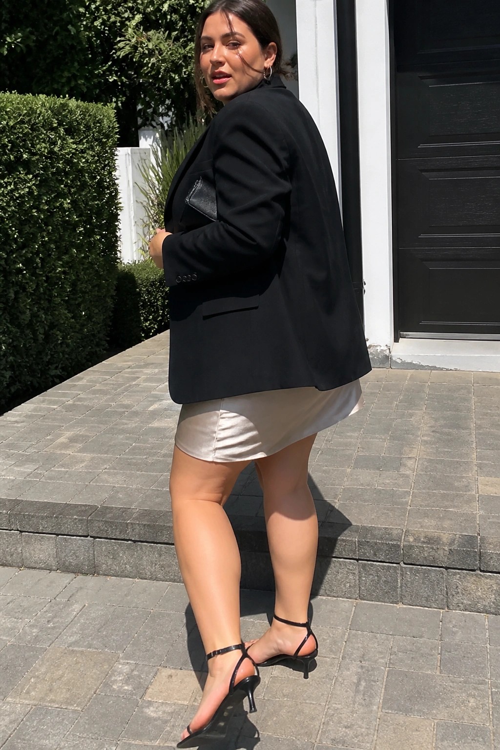 Plus size woman in oversized black blazer over white satin mini skirt with black strappy heels standing on stone steps.