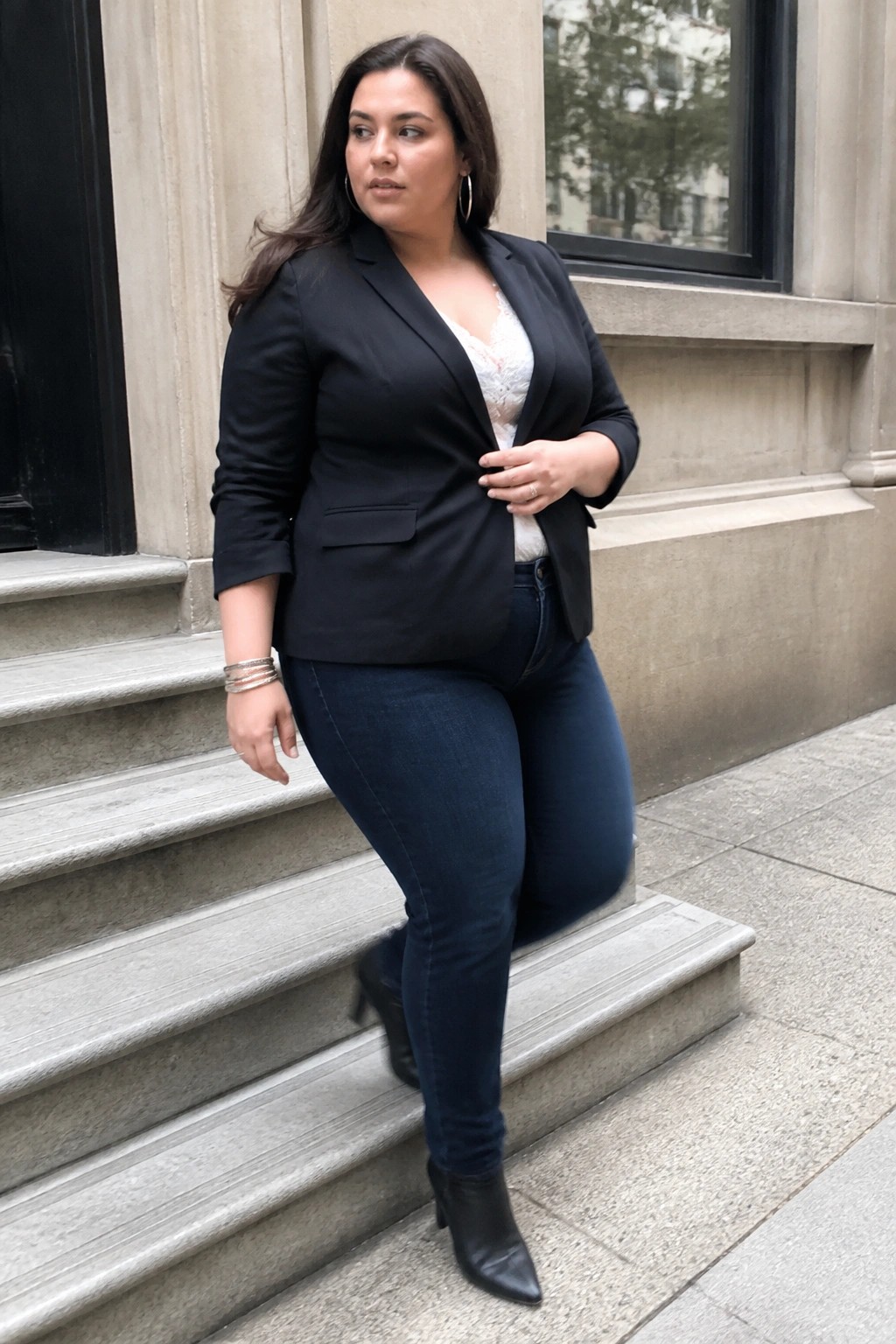 Plus size woman in black blazer over white lace top paired with dark blue jeans and black ankle boots on stone steps