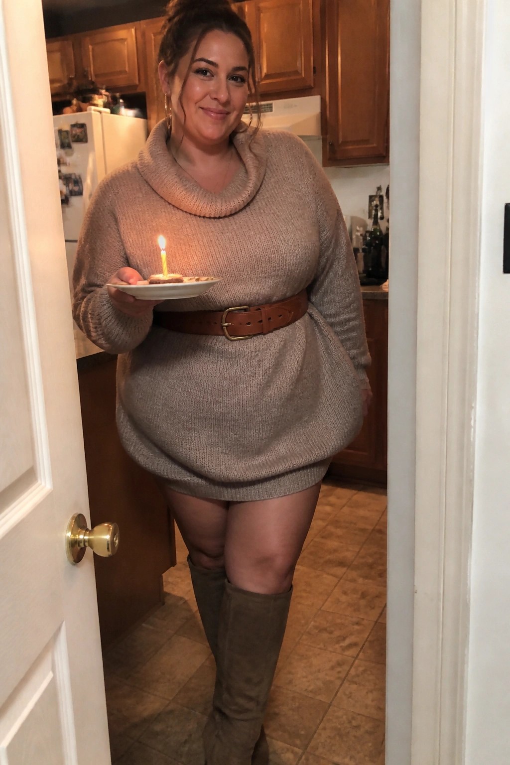 Woman in beige turtleneck sweater dress holding plate with lit candle in kitchen