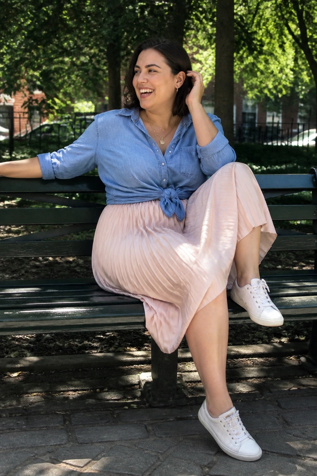 Smiling woman in tied blue denim shirt and pink pleated skirt on park bench