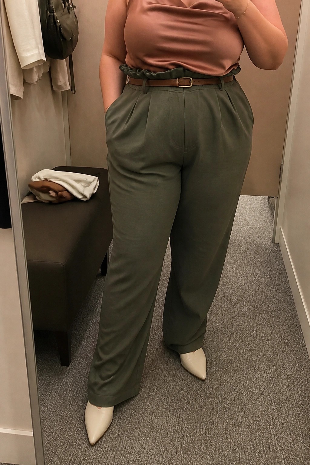 Woman in sage green pleated trousers, pink satin top, brown belt, white heels.
