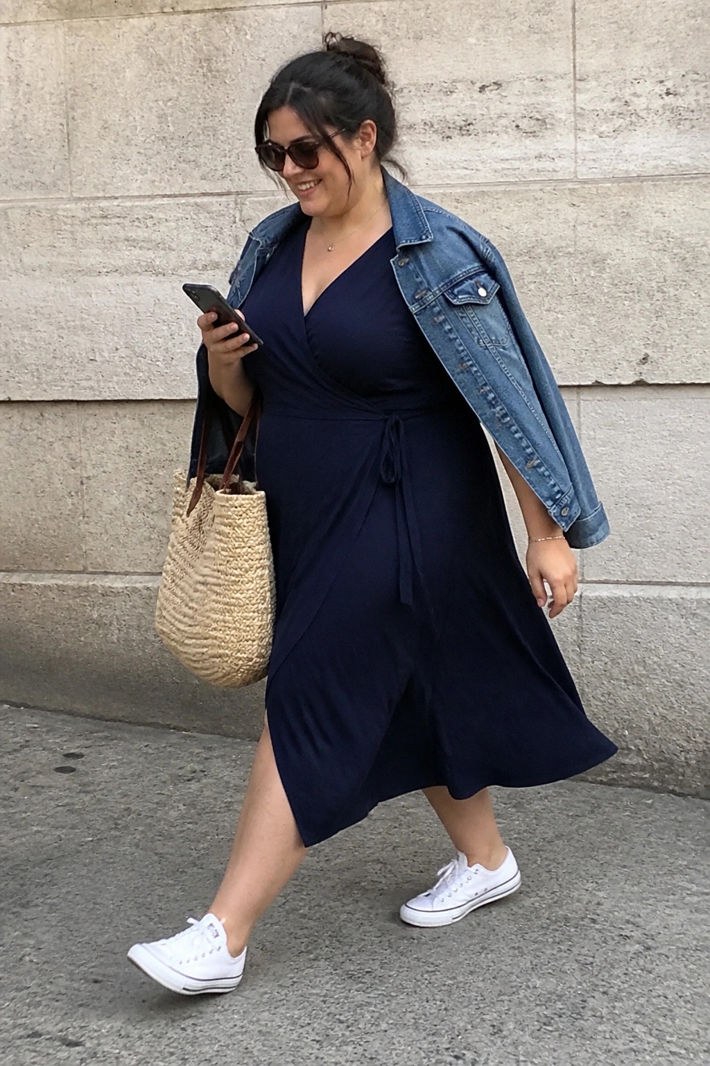 Smiling woman in navy wrap dress and denim jacket walks with straw bag and phone.