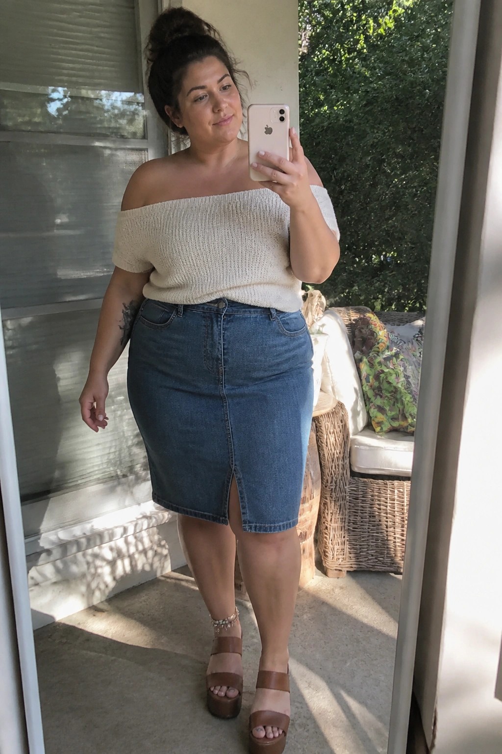 Woman in beige off-shoulder knit top and blue denim skirt taking iPhone selfie on porch.