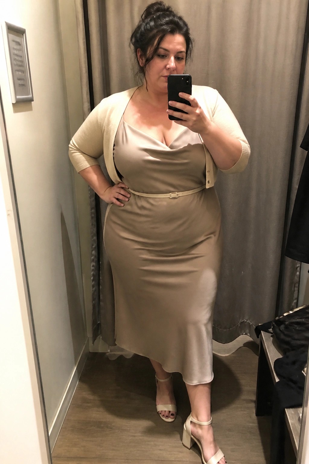 Plus size woman wearing a beige satin midi dress with cowl neck, cream cardigan, thin beige belt, and matching block heel sandals while taking a mirror selfie in a fitting room.
