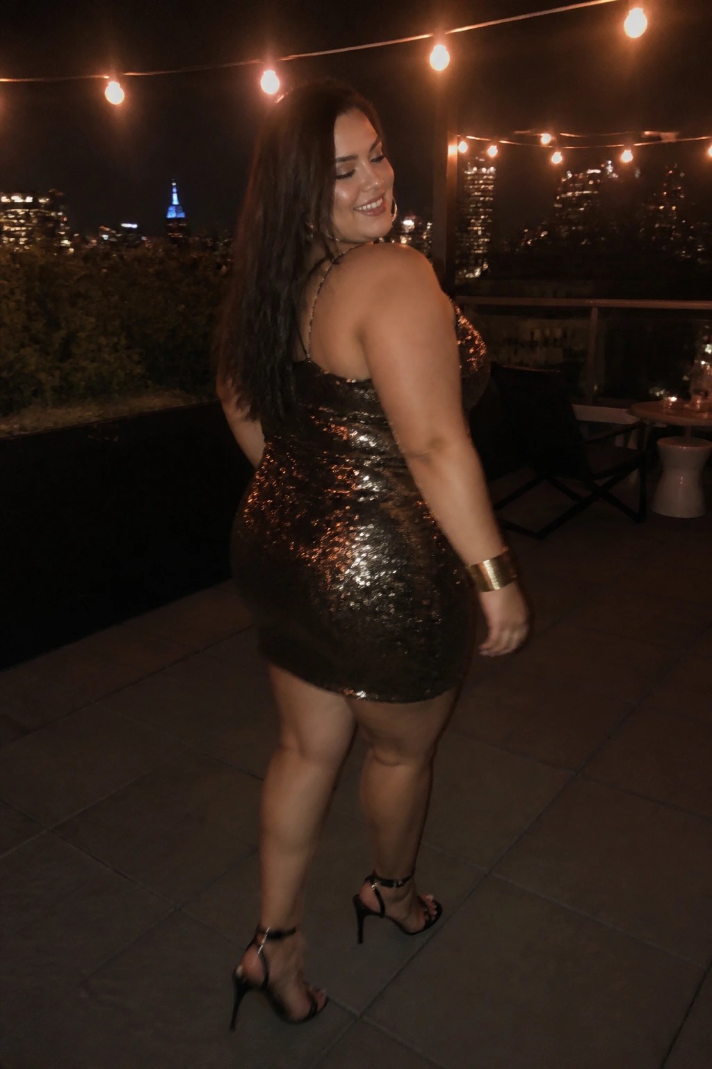 Woman in sequined mini dress posing on nighttime rooftop with city lights.