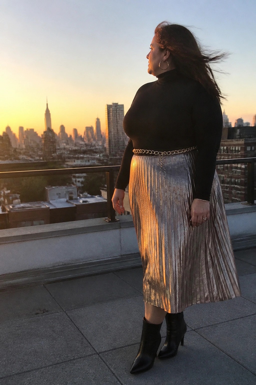 Woman in black turtleneck and metallic silver pleated skirt on NYC rooftop at sunset