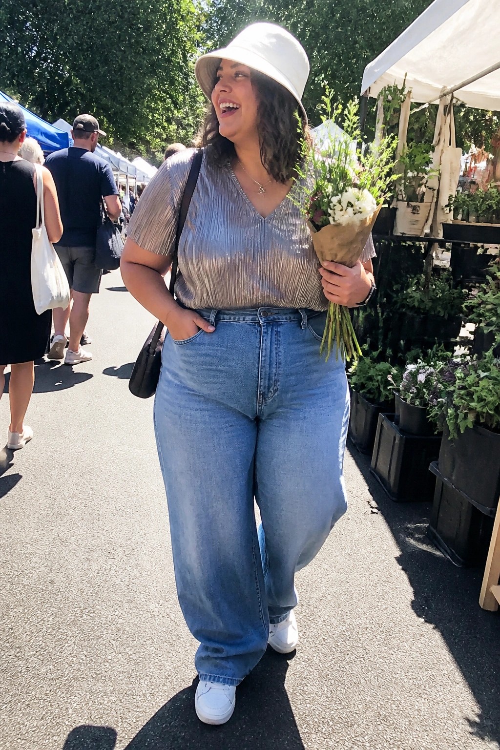 Smiling woman in white hat and shiny top holds flowers at sunny market