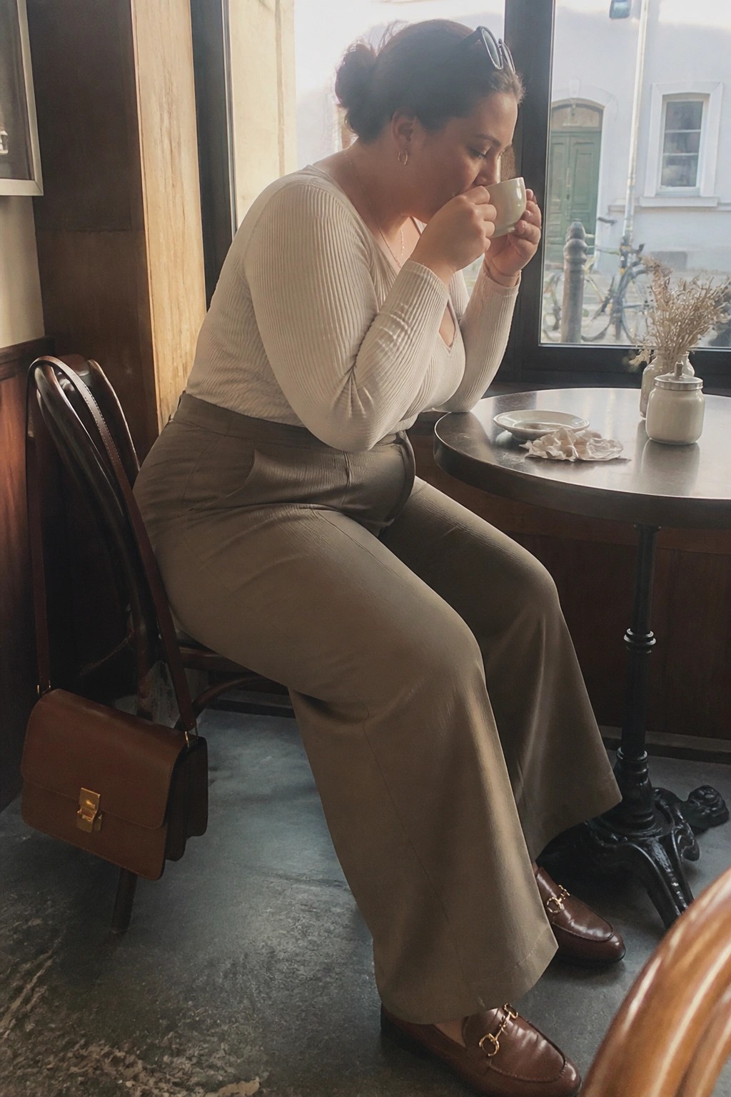 Woman in cream ribbed V-neck top and taupe wide-leg pants seated at a cafe table holding a coffee cup with brown leather loafers and matching crossbody bag visible.