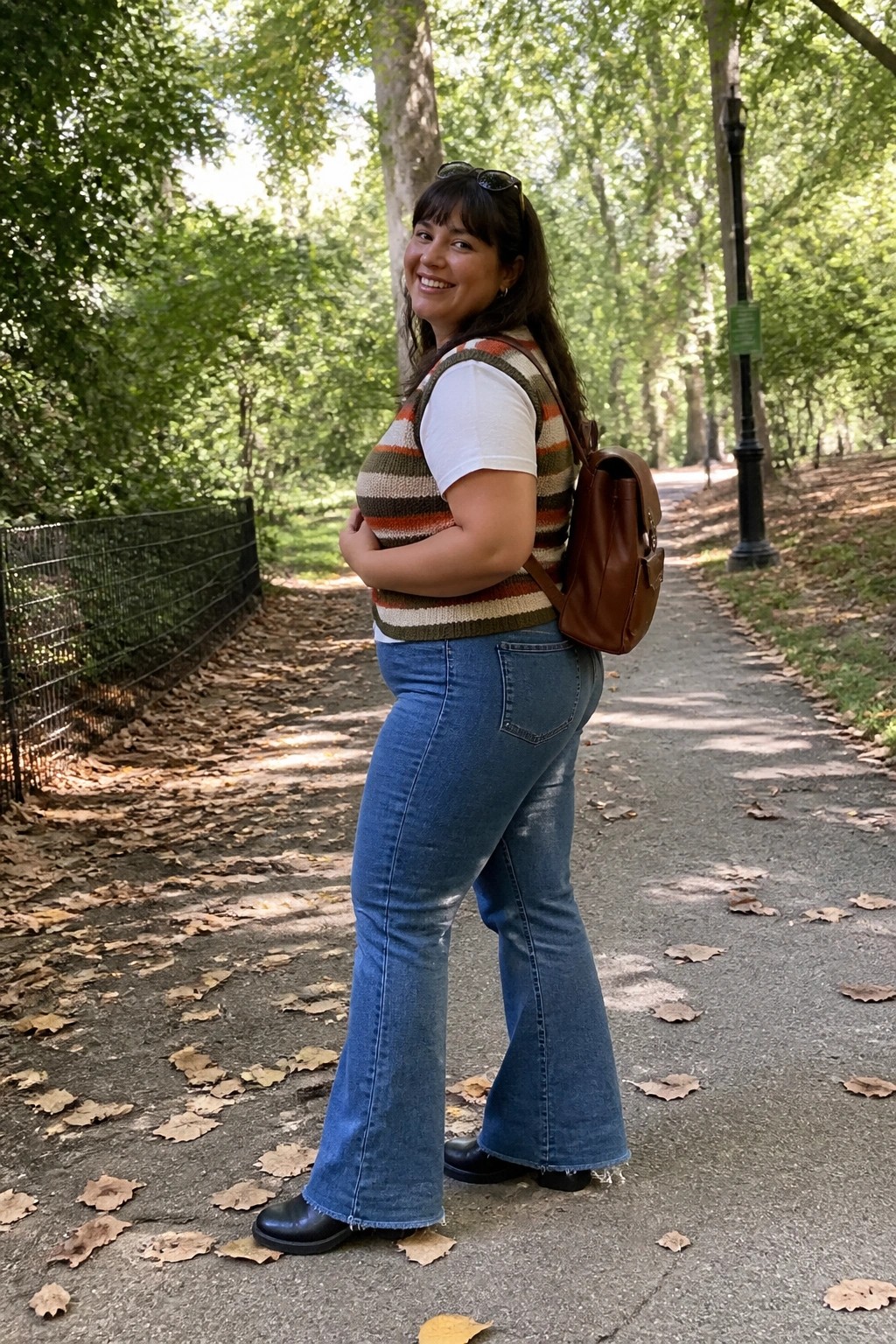 Plus size woman wearing blue flare jeans with a multicolored striped knit vest over a white t-shirt, brown leather backpack, and black shoes standing on a path.