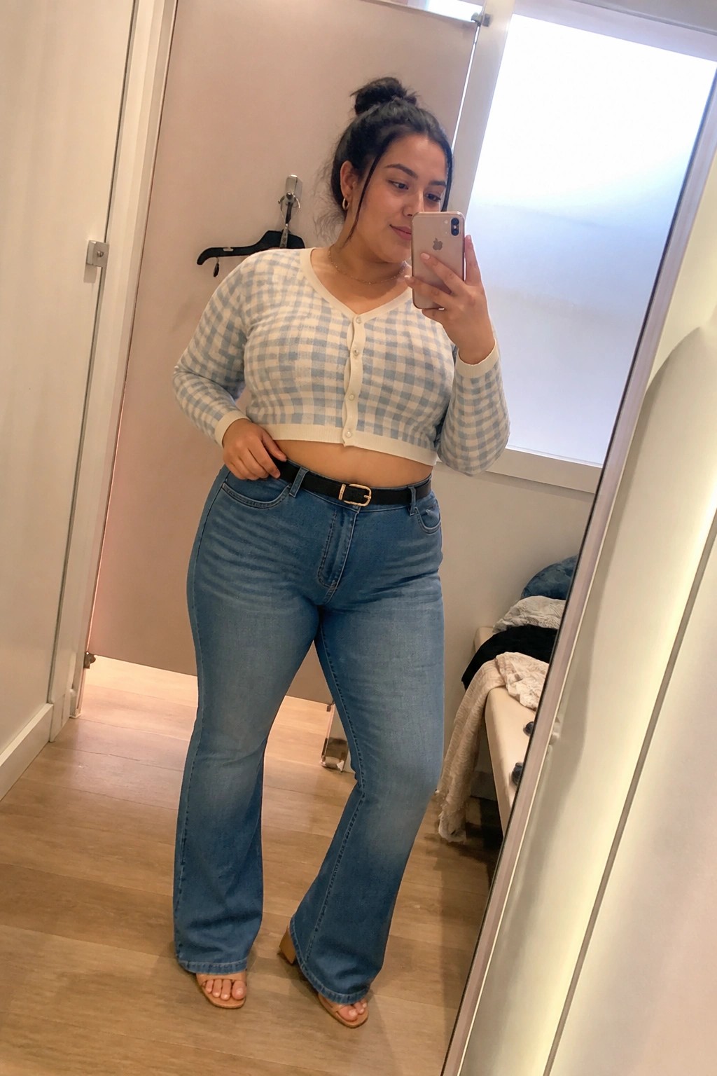 Woman in blue gingham crop top and flared jeans taking mirror selfie with phone