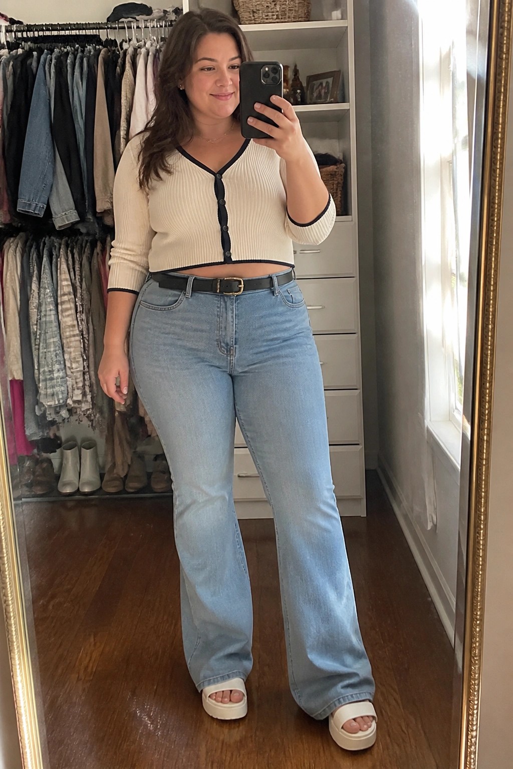 Woman in cream cardigan and light blue jeans taking mirror selfie in closet