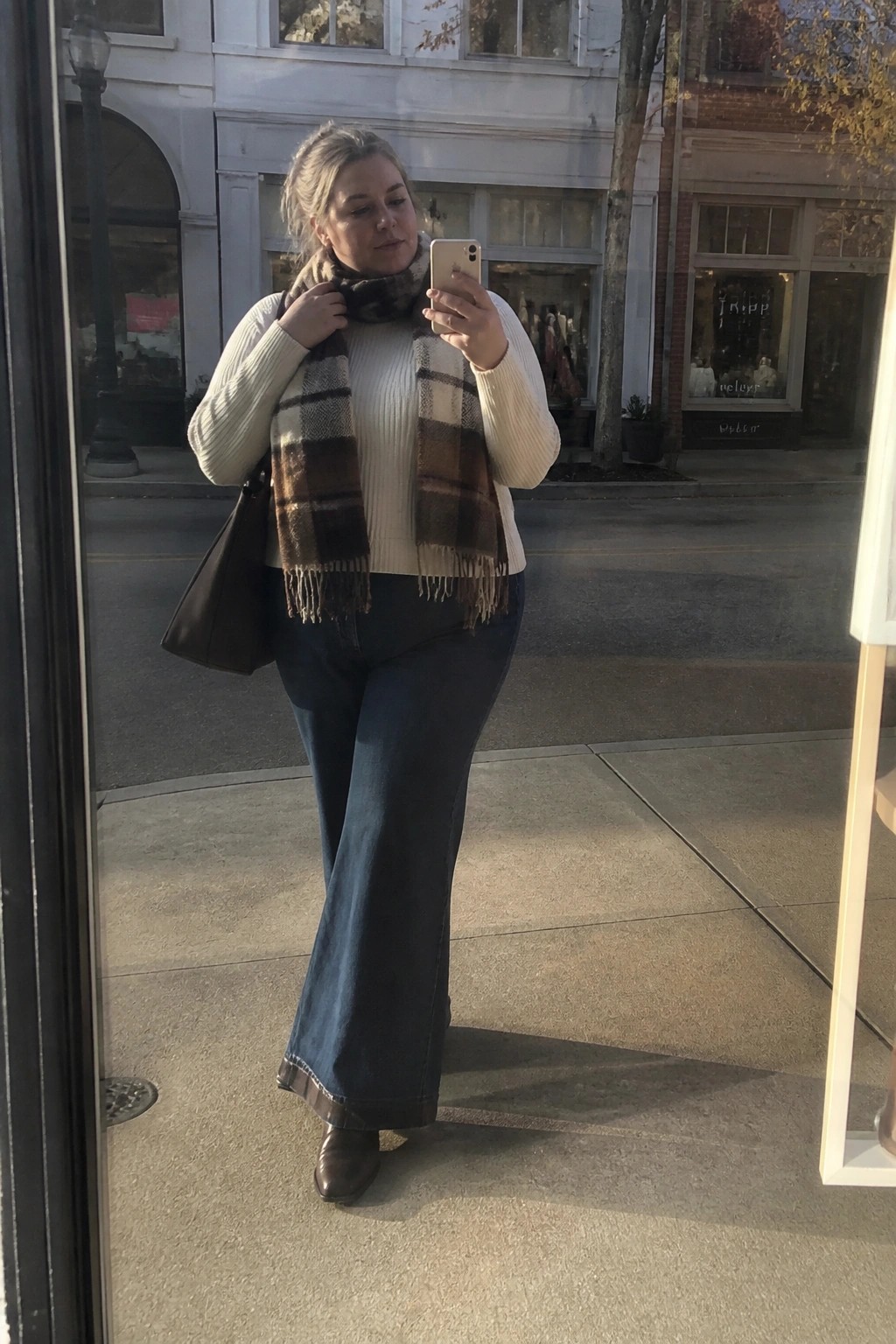 Woman in white sweater and plaid scarf taking mirror selfie on city sidewalk