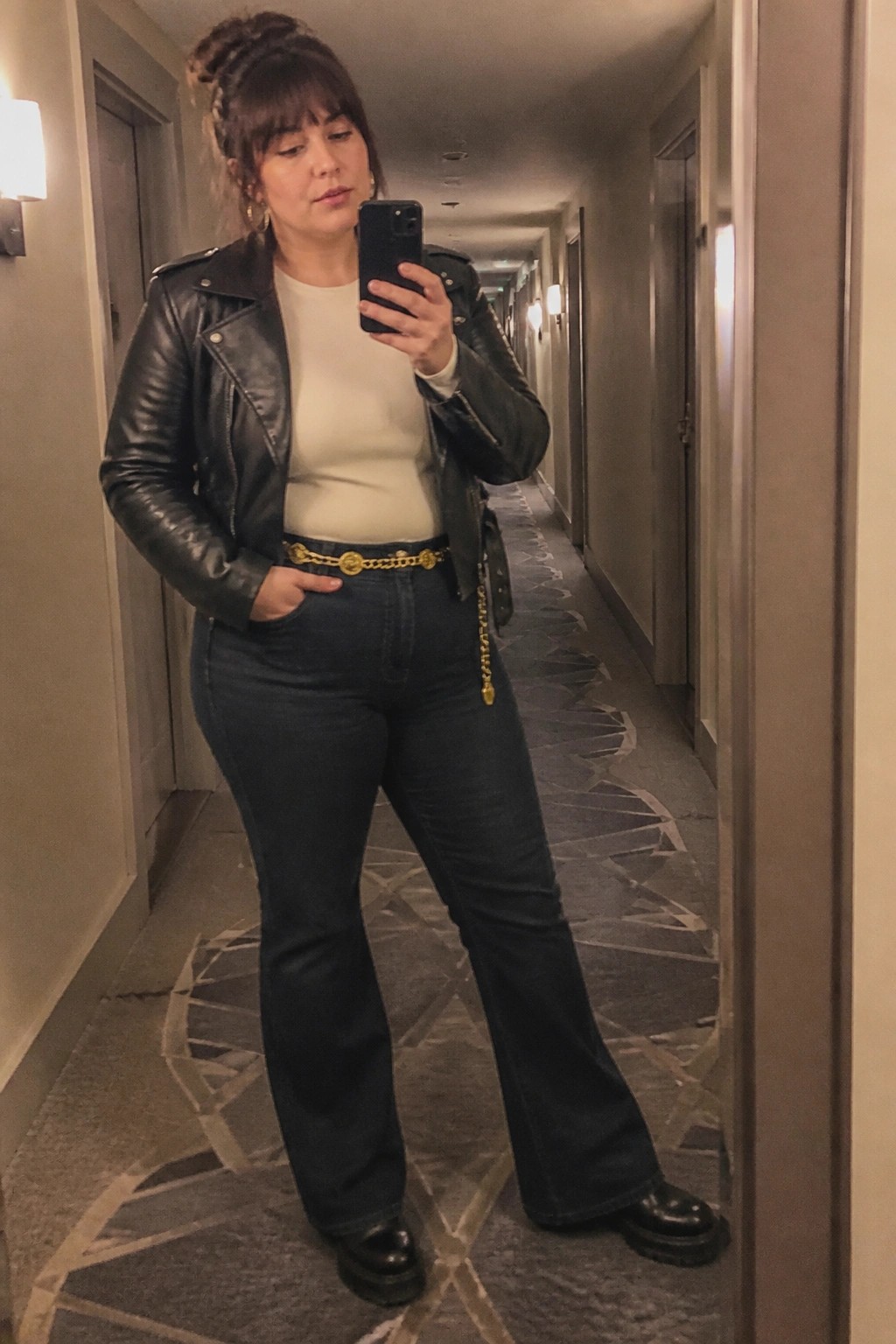 Woman in black leather jacket and jeans taking mirror selfie in hallway