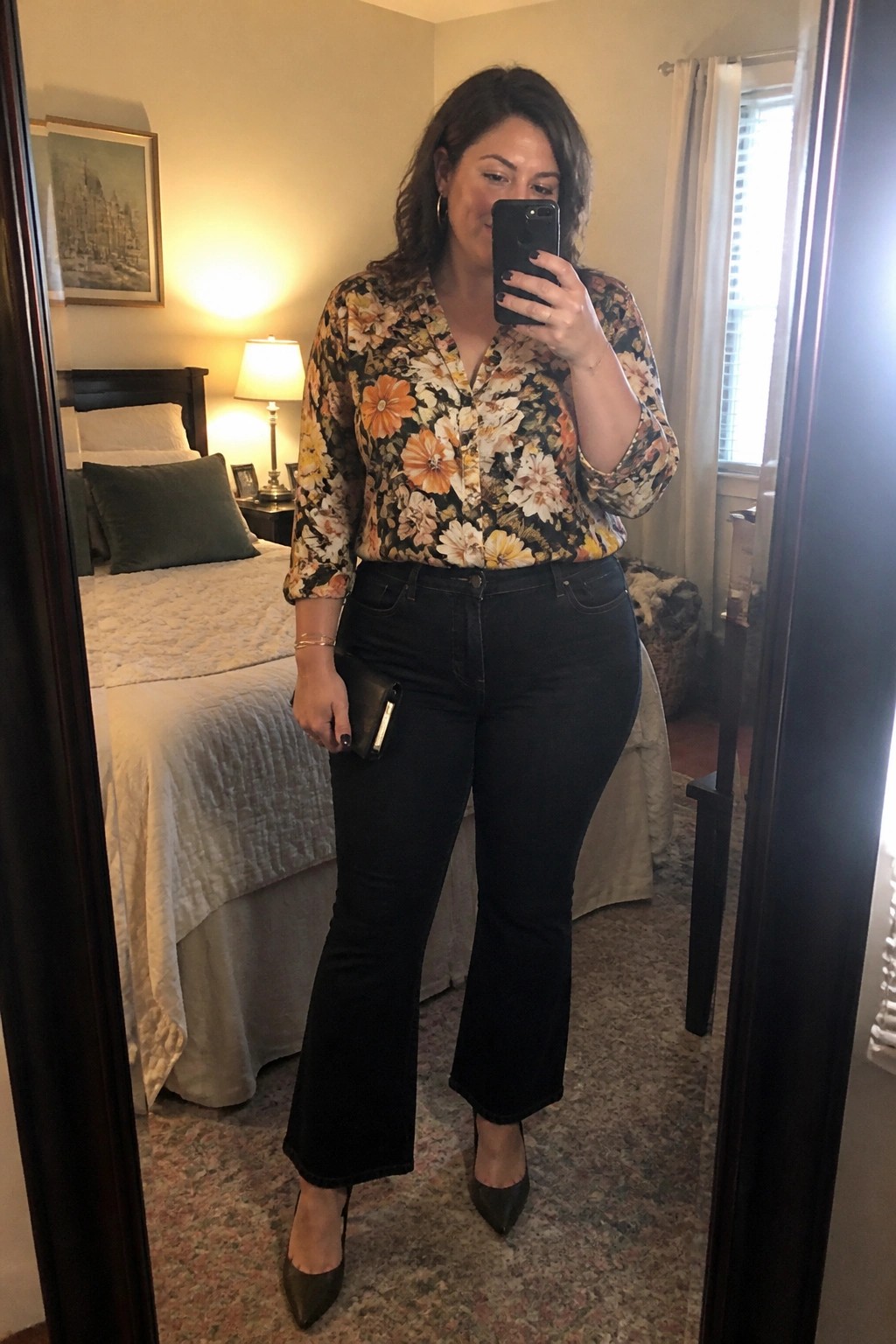 Woman wearing a black floral print blouse tucked into black flare jeans with dark pointed toe heels while holding a phone for a mirror selfie.