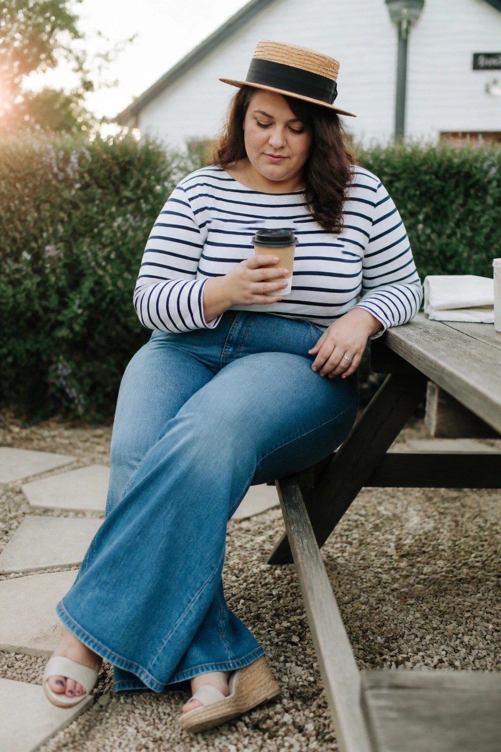 Woman in striped shirt and straw hat holds coffee at picnic table