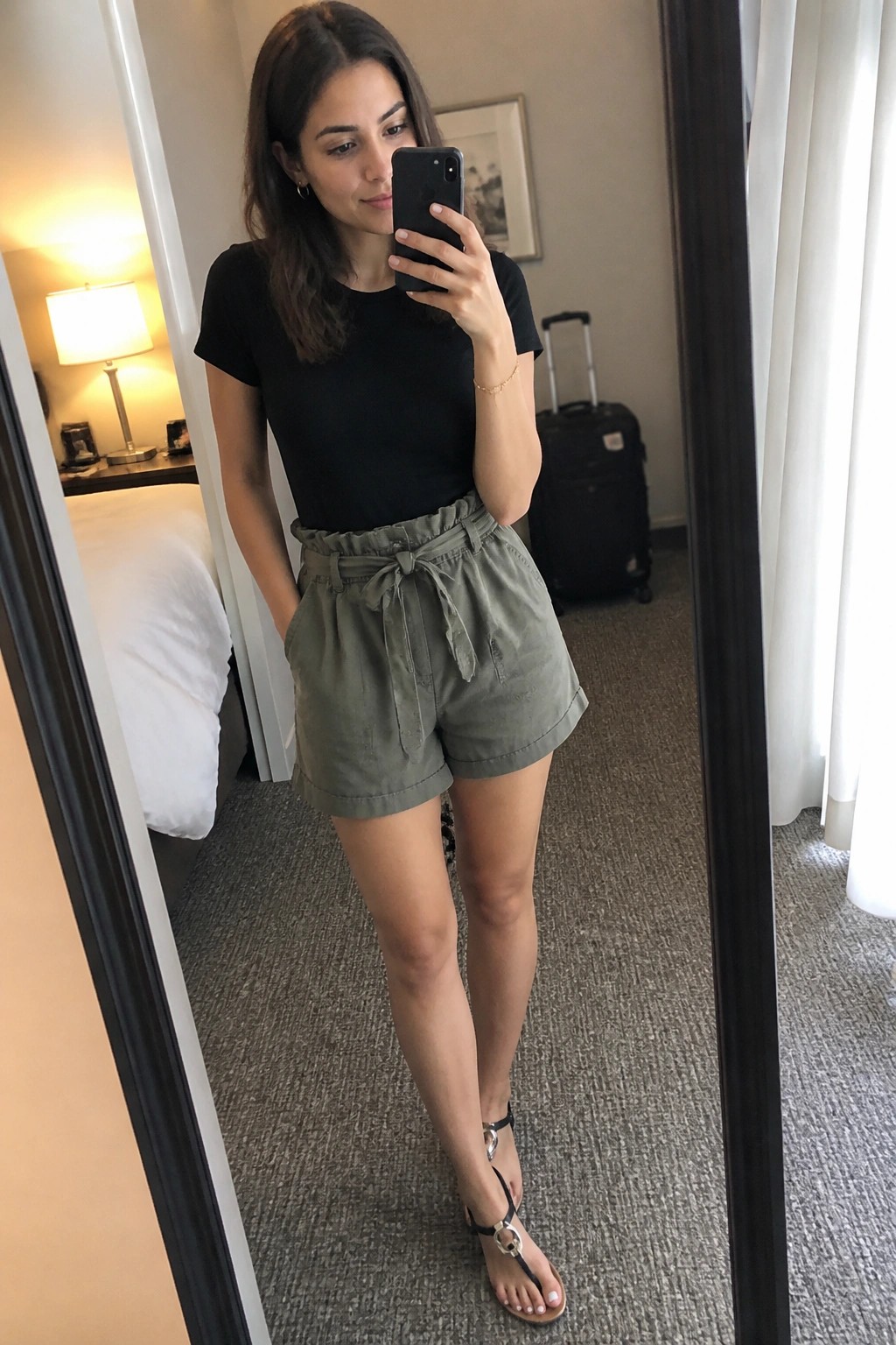 Woman in black tee and olive tie-waist shorts taking mirror selfie in hotel room