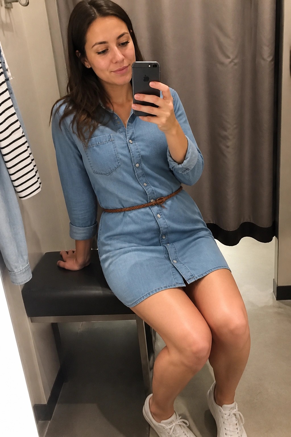Woman in blue denim shirt dress taking mirror selfie on black bench