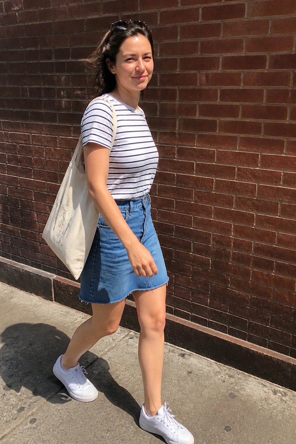 Woman in striped shirt and denim skirt with tote bag by brick wall