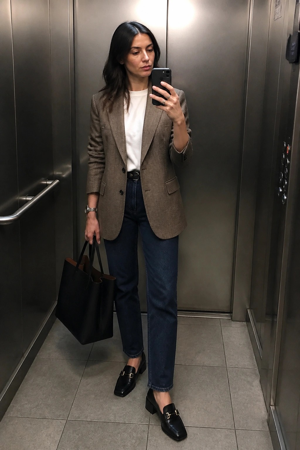 Woman in brown blazer and jeans taking mirror selfie in elevator with tote bag