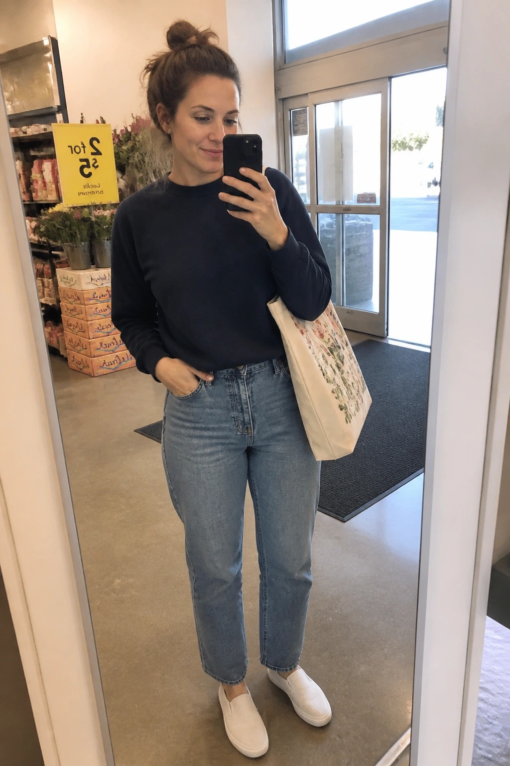 Woman in navy sweatshirt and jeans taking mirror selfie with floral tote bag