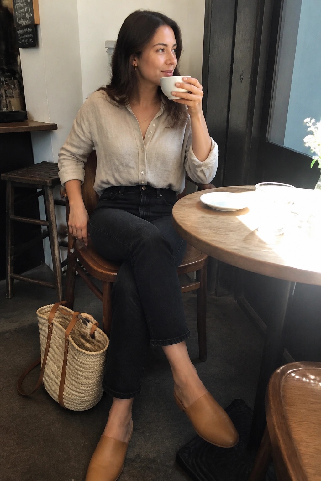 Woman in beige linen shirt sips from cup at wooden cafe table with woven bag.