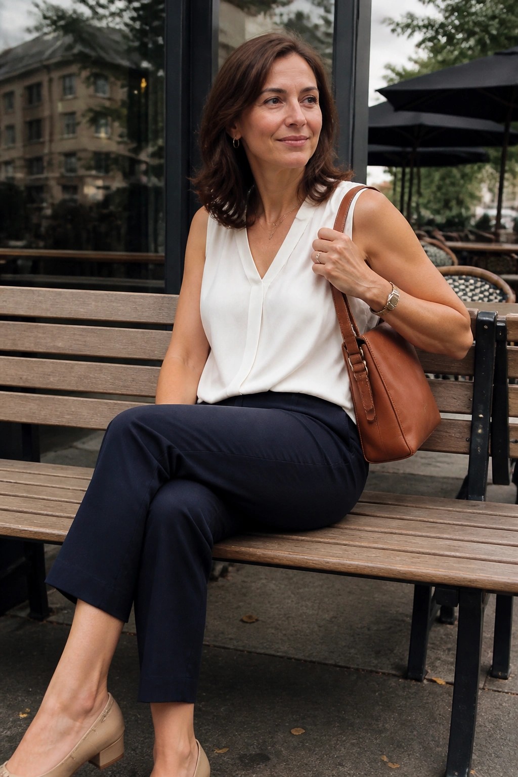 Woman seated on a bench wearing a white sleeveless v neck blouse tucked into navy cropped trousers with beige low block heel shoes and carrying a brown leather shoulder bag.