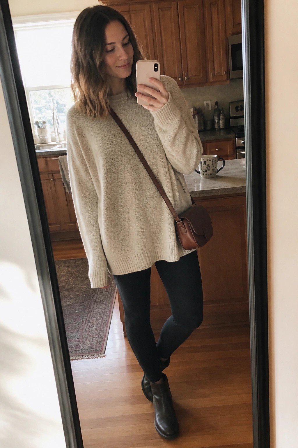 Woman in cream oversized sweater taking mirror selfie in kitchen with brown bag