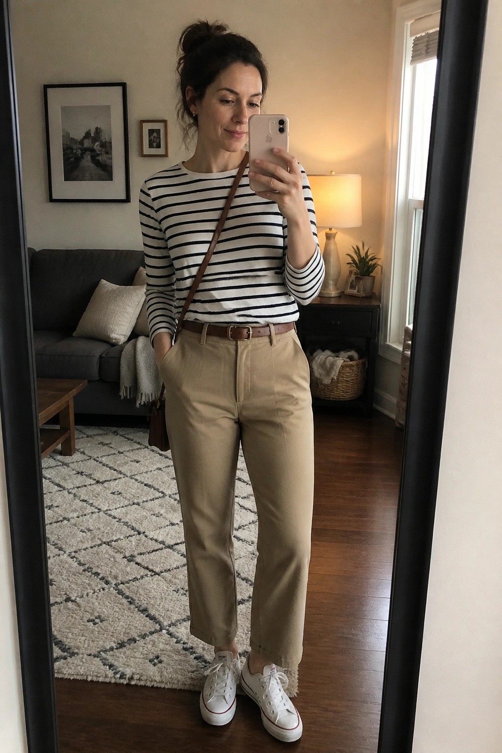 Woman in striped shirt and khaki pants taking mirror selfie in living room
