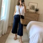 Woman in white sleeveless top and navy culottes taking bedroom mirror selfie with woven bag