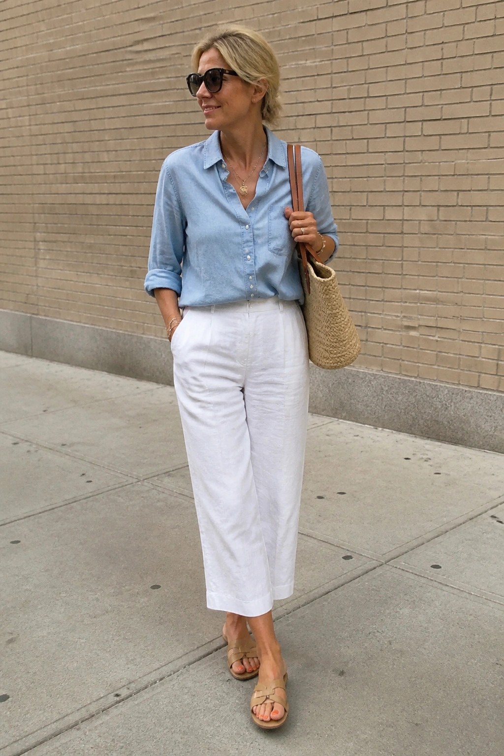 Woman in light blue shirt and white pants holding woven tote on sidewalk