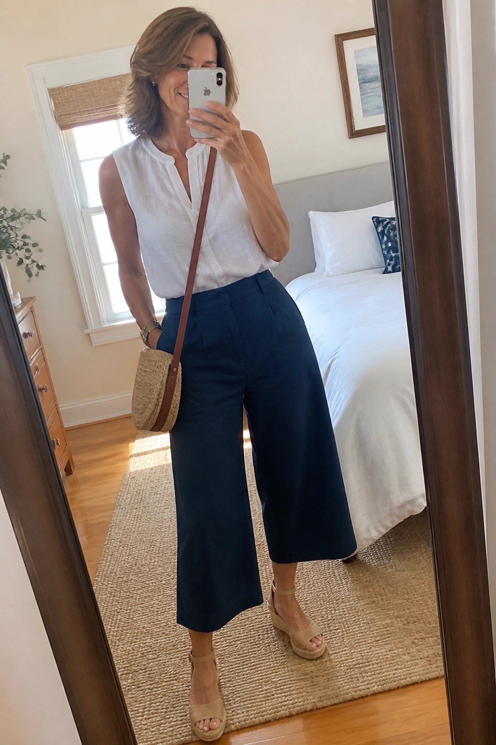 Woman in white sleeveless top and navy culottes taking bedroom mirror selfie with woven bag
