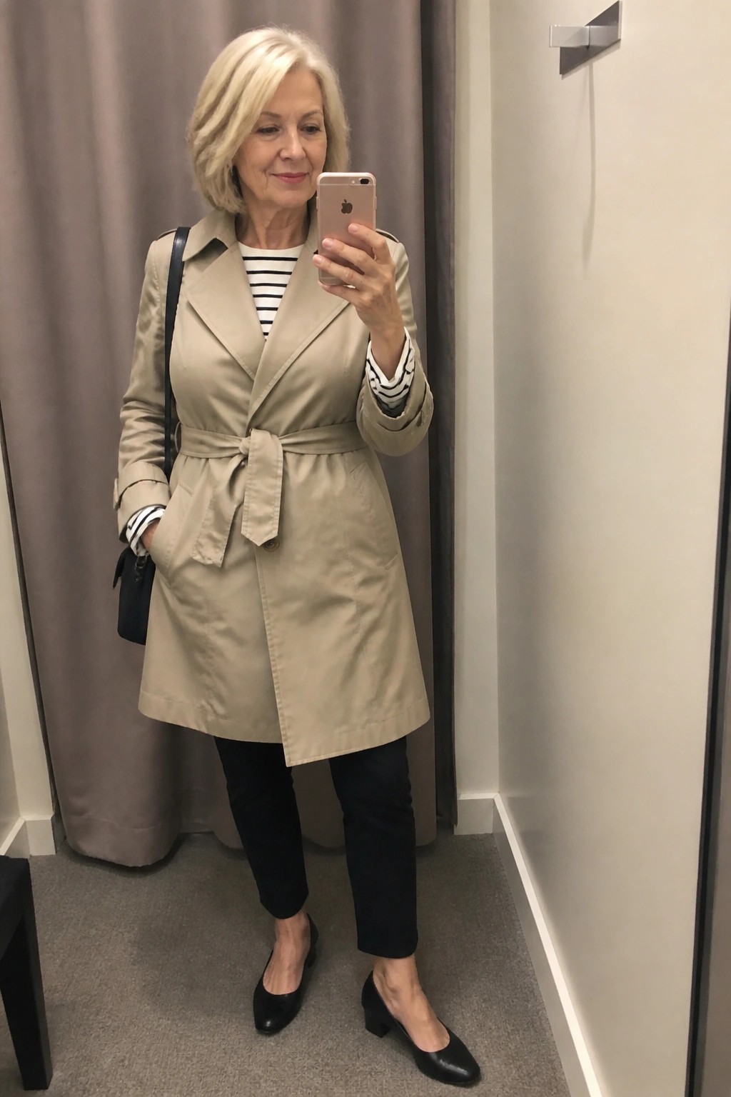 Woman in beige trench coat belted at the waist over a black and white striped shirt with black pants and low black heels holding a phone for a mirror selfie