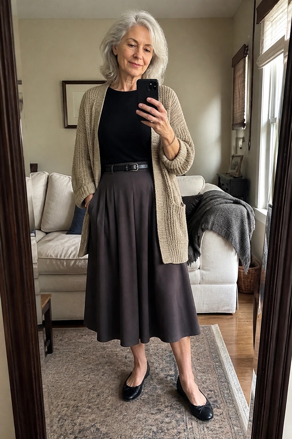 A woman with gray hair wearing a beige open cardigan over a black top, dark gray pleated midi skirt with black belt, and black ballet flats, taking a mirror selfie.