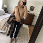 Woman with gray hair in beige turtleneck, jeans, and boots holding plaid coat for mirror selfie