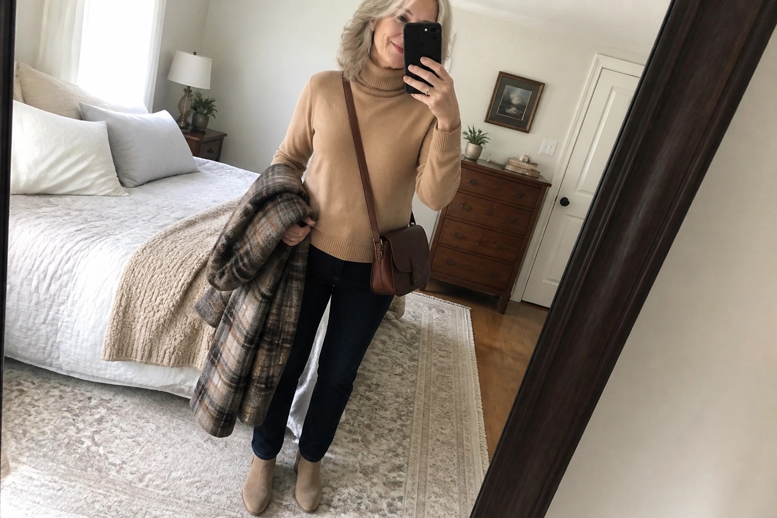 Woman with gray hair in beige turtleneck, jeans, and boots holding plaid coat for mirror selfie