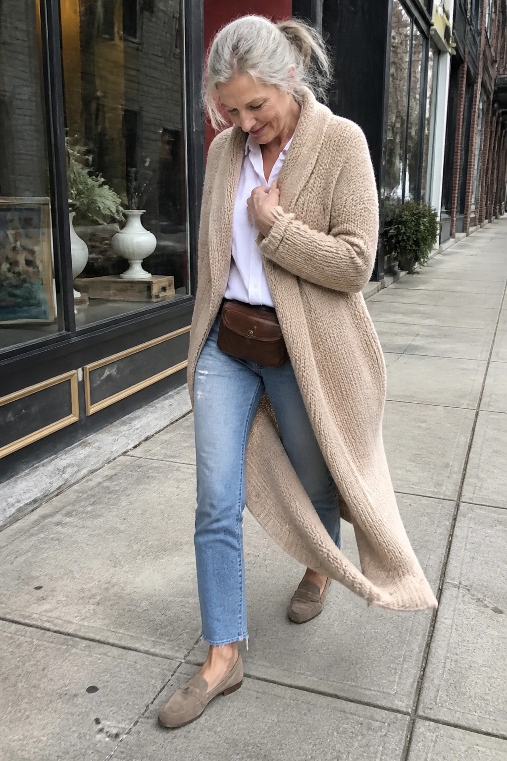 Woman in beige knit cardigan, white shirt, and jeans on urban sidewalk