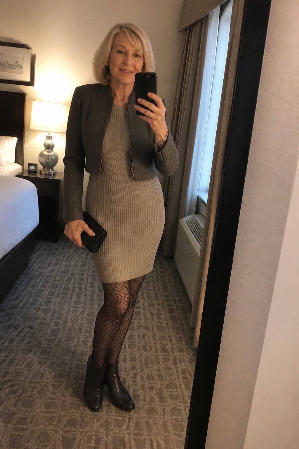 Older woman in beige knit dress and gray jacket taking mirror selfie