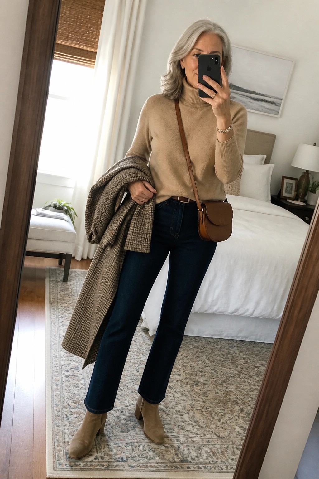 Woman with gray hair in beige turtleneck, jeans, and boots holding plaid coat for mirror selfie