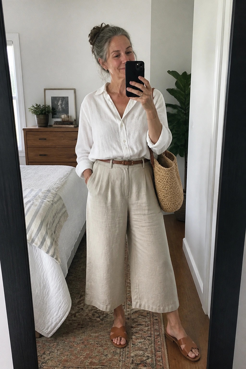 Woman in white shirt and beige pants taking mirror selfie in bedroom