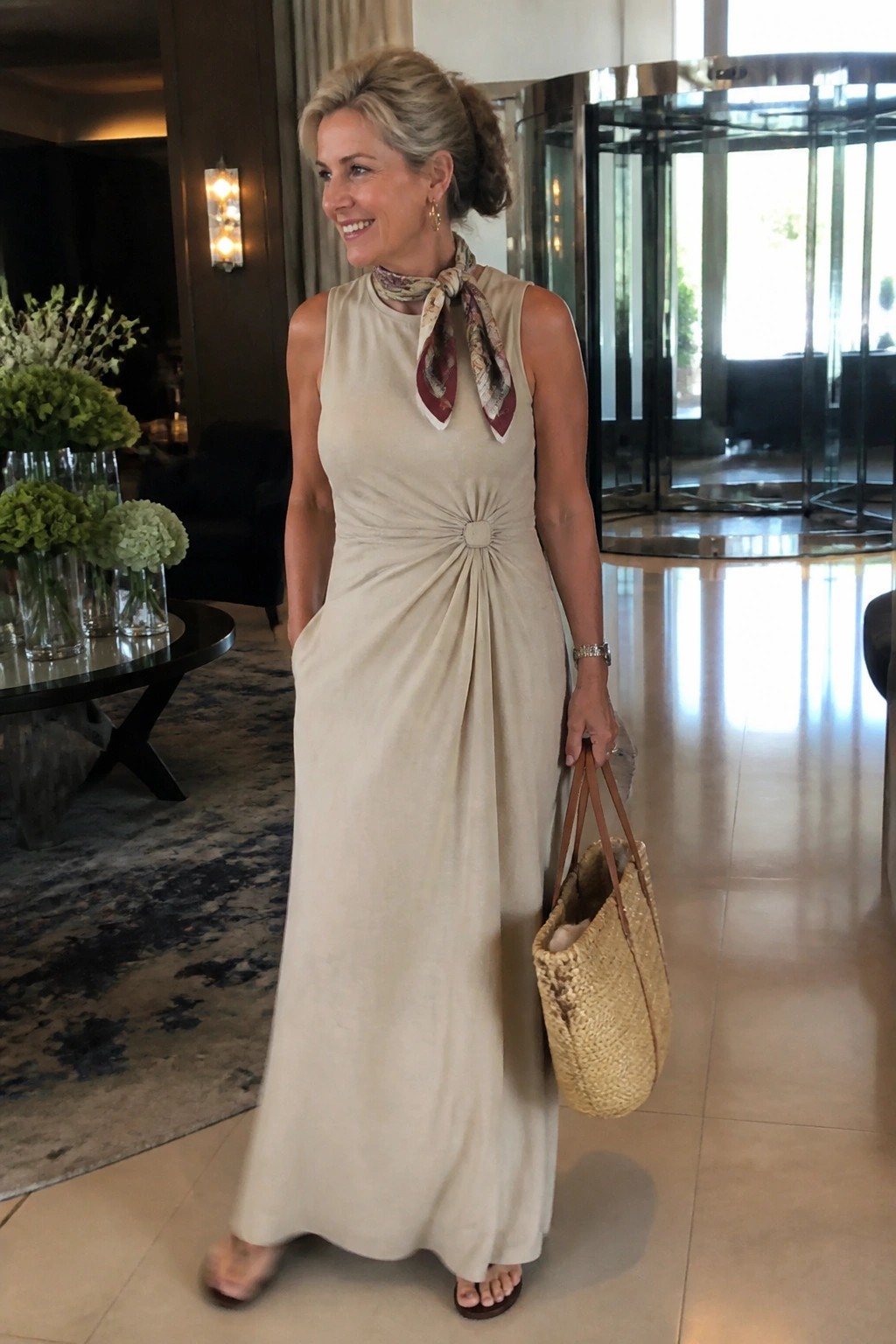 Woman in beige ruched maxi dress with scarf holding straw tote in lobby