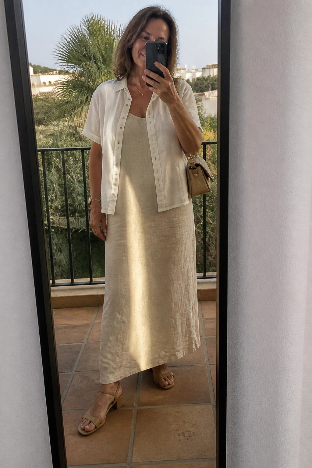 Woman in white shirt over beige dress taking balcony mirror selfie with phone
